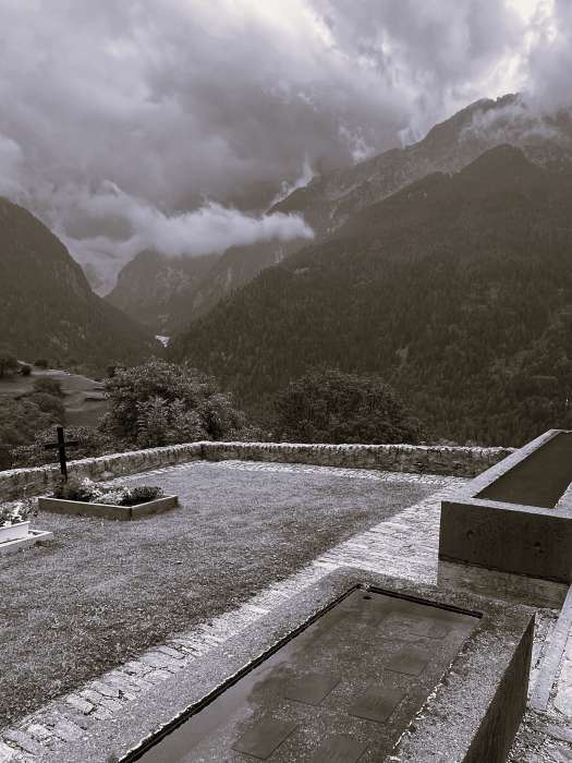 Aufnahme mit Grab und Grabkreuz, graue Wolken und Bergen schwarz-weiss Fotografie Thomas Pfister
